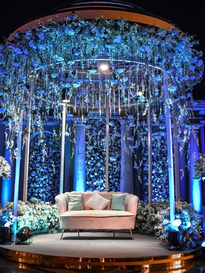 A stunning stage setup with an 'Enchanted Blue' theme. A canopy of blue flowers hangs over the seating area, which is set against a backdrop of a thousand more blue blooms, all illuminated to create a magical, monochromatic world.
