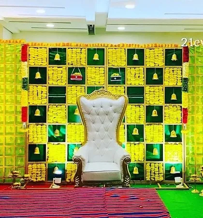 A wide-angle view of a traditional baby shower stage. The backdrop features a checkerboard of betel leaves and marigolds, with a grand white throne as the centerpiece.