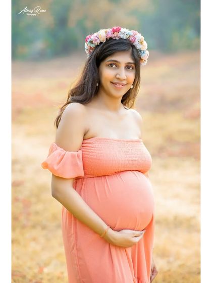A beautiful, direct portrait of the mom-to-be. Wearing a floral crown and an off-the-shoulder peach gown, her happy expression and radiant glow are the central focus of the image.