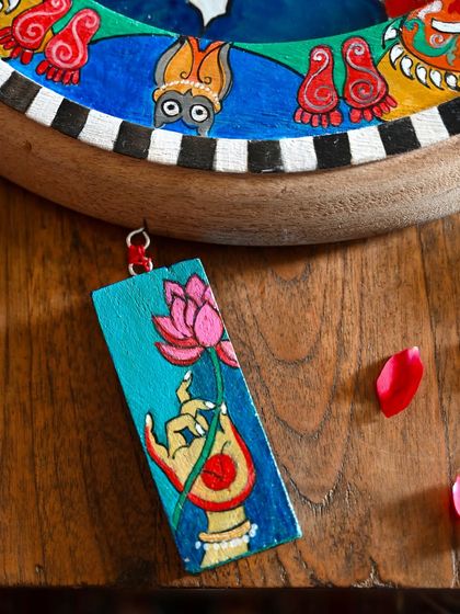A close-up of one of the hand-painted wooden charms hanging from the Laxmi frame. This small detail, showing a hand holding a lotus, adds another layer of symbolic meaning and handcrafted beauty to the piece.