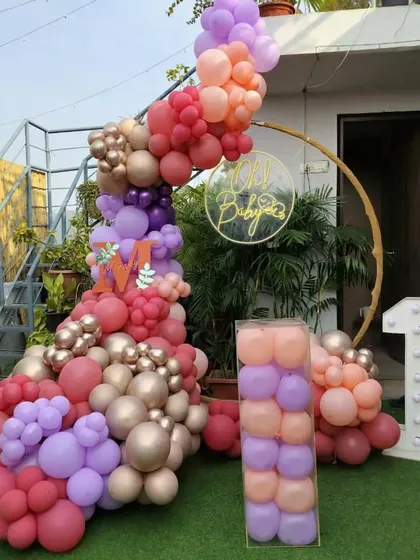 An outdoor first birthday setup on a grassy lawn. It features a gold ring backdrop with a neon sign, a large balloon arrangement in shades of pink and purple, and a light-up number '1'.