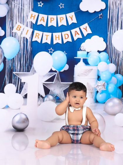 A full view of the blue and silver birthday setup, with the baby curiously looking at his cake. This shot showcases the entire festive scene, from the balloons to the "Happy Birthday" banner.