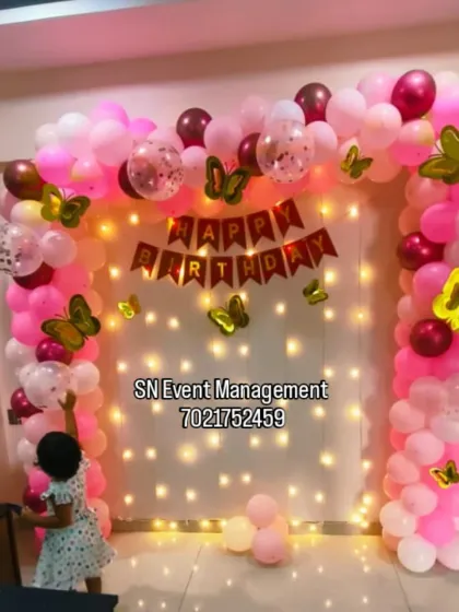 A sweet butterfly-themed decoration for a first birthday at home. The pink balloon arch is set against a curtain with fairy lights, creating a magical glow for the little one.