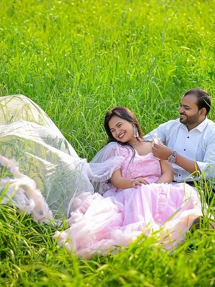 A sweet and candid moment captured in a lush green field. The groom helps the bride with her necklace as she lies back on a flowing veil, creating a dreamy and intimate pre-wedding portrait.