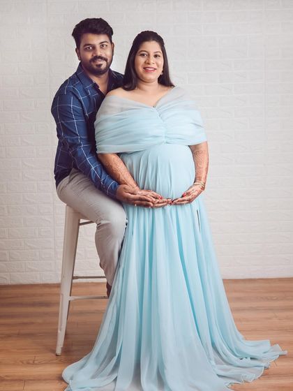 A classic seated pose for a couple's maternity shoot. The light blue off-the-shoulder gown looks absolutely dreamy against the simple white brick background.