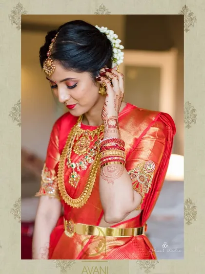 A bride getting ready, wearing a red Kanchipuram saree. Her custom blouse is adorned with heavy gold embroidery on the sleeves, perfect for her muhurtham.