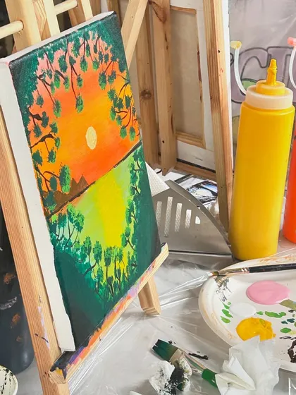 A close-up of a sunset painting in progress, with bottles of colorful paint ready to be used.