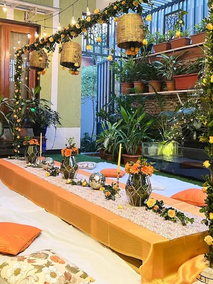 An intimate dinner celebration at home featuring a beautiful low-seating arrangement. The decor includes an orange and yellow color scheme, a floral arch, and hanging basket lanterns.