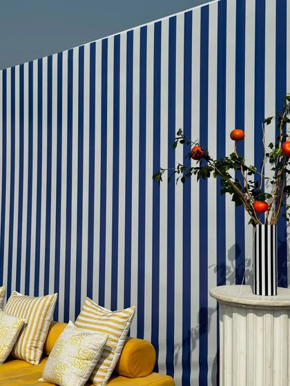 A stylish seating area with a mustard yellow sofa and custom cushions. The backdrop is a bold blue-and-white striped wall, accented by a pedestal with a vase holding branches of pomegranates.