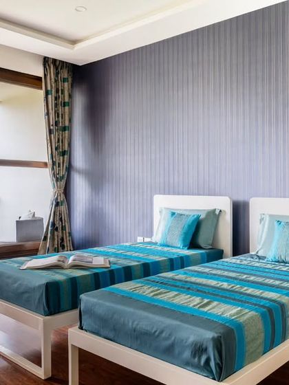 For this children's or guest bedroom, we used two single beds to maximize flexibility. The deep blue accent wall and bright turquoise bedding add a playful yet sophisticated pop of color, while the large window connects the room to the lush greenery outside.