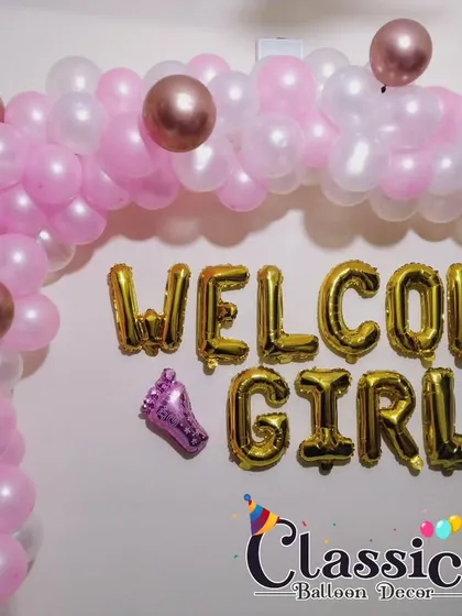 A 'Welcome Girl' decoration featuring a pink and rose gold balloon arch with matching foil letters.