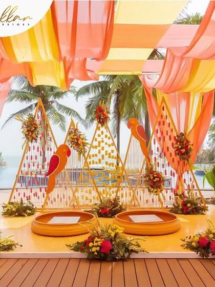A vibrant poolside Haldi setup featuring parrot-themed decor and urlis. The bright orange and yellow drapes create a tropical and festive atmosphere.