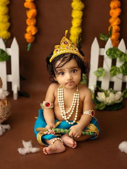 An adorable solo shot of a baby dressed as Krishna, seated among props like golden matkis and a small flute.
