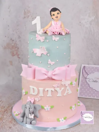 A sweet two-tier cake for a first birthday, combining butterflies, bows, florals, and a cute baby elephant.