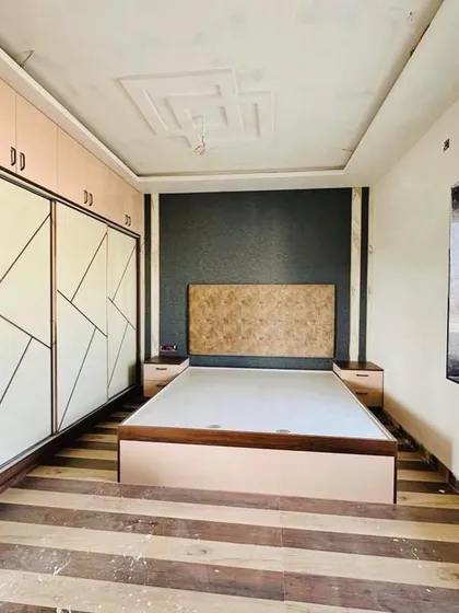 This bedroom in a lengthy room features a lacquered glass sliding wardrobe with golden T-strips. The minimal cushion headboard is set against a dark highlighter wall to create a stunning focal point.