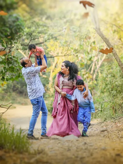 A family of four shares a joyful, candid moment on a sun-dappled forest path. The father lifts their youngest child high in the air, showcasing the playful energy and connection I capture during my outdoor family sessions.