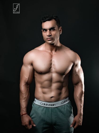 A powerful, direct-to-camera stance that showcases a full and well-developed chest and shoulders. The simple grey trousers and dark background keep the focus entirely on the upper body physique.