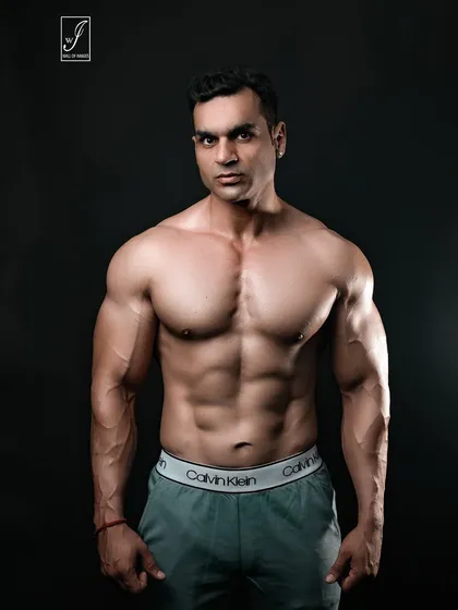 A powerful, direct-to-camera stance that showcases a full and well-developed chest and shoulders. The simple grey trousers and dark background keep the focus entirely on the upper body physique.