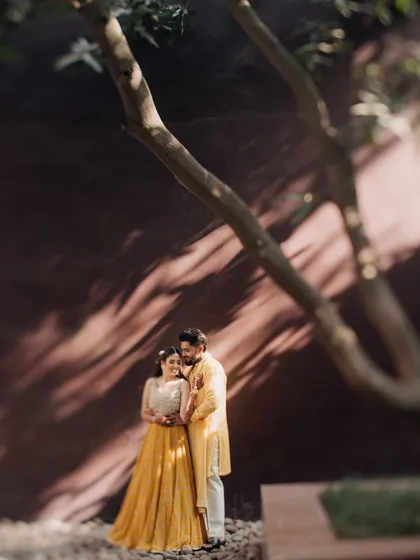 An artistic portrait using natural light and shadows. The tree's shadow creates a beautiful, dappled effect on the wall, framing the couple in a serene and romantic pre-wedding moment.