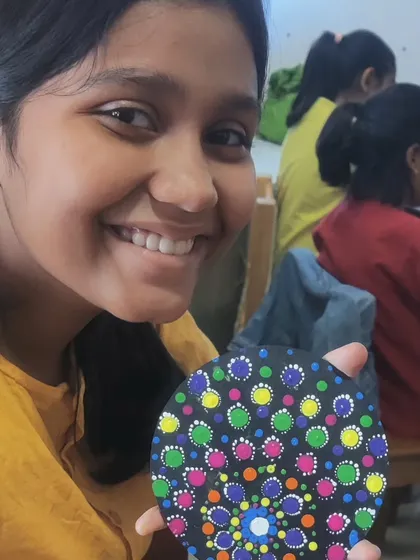 A smiling student shows her colourful dot art coaster. The bright pattern of green, yellow, and pink dots on the black background is cheerful and eye-catching.