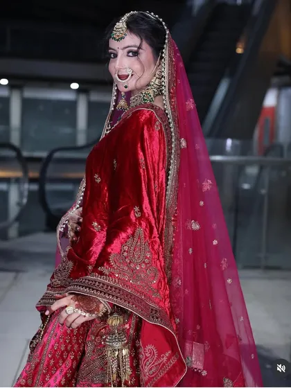 A side view of a bride in a rich red velvet lehenga, showing the detailed embroidery and the flow of the sheer pink dupatta.