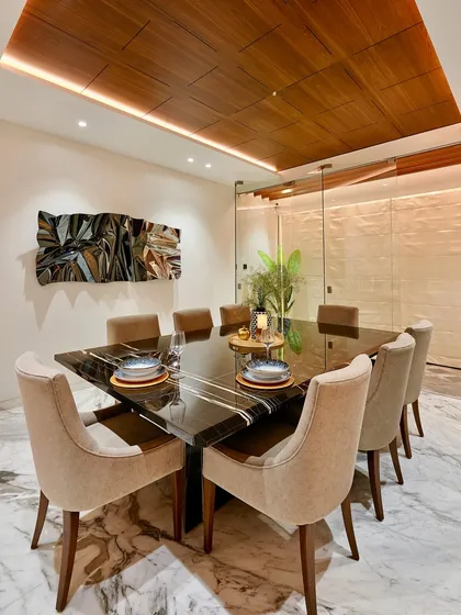 A perspective shot of a formal dining area. The square black marble table, seating eight, is positioned under a coffered wood ceiling with perimeter lighting, creating an intimate yet grand setting for meals.