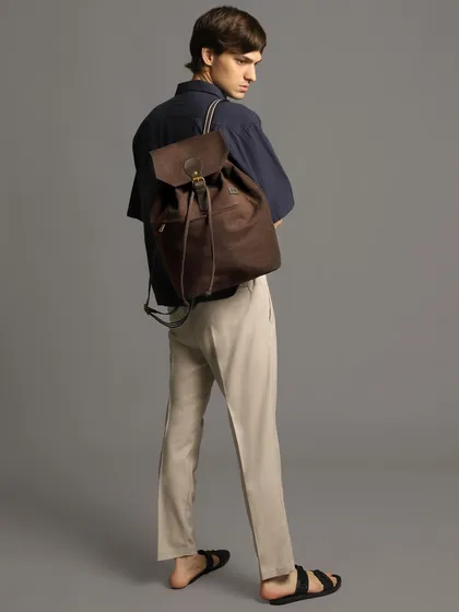 An accessory-focused shot from a men's editorial. The model's relaxed pose highlights a suede backpack, showing how the right bag can complete a casual, stylish look.