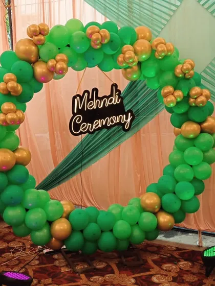 A close-up of a Mehndi ceremony backdrop I designed. This features a full ring of green and gold balloons, which are classic Mehndi colors, with a custom 'Mehndi Ceremony' sign in the center. It’s a perfect photo booth spot.