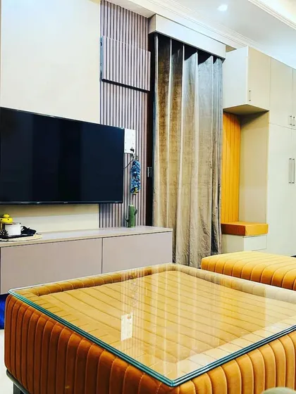 A different angle of the Jasola living room, highlighting the vibrant orange upholstered ottomans that double as a coffee table. This multi-functional furniture piece adds a playful yet practical element to the space, contrasting beautifully with the neutral tones of the TV unit.