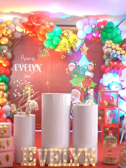 A festive Christmas-themed first birthday for Princess Evelyn. The red backdrop is adorned with a colorful balloon arch featuring bell and star foil balloons, creating a joyful holiday atmosphere.