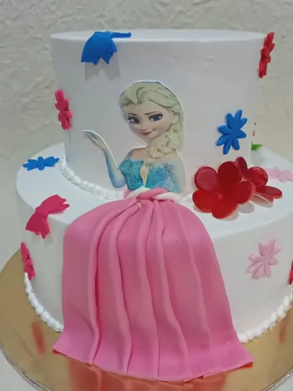A two-tiered cake featuring an Elsa motif. The design is complemented by a flowing pink fondant "dress" that drapes down the side.