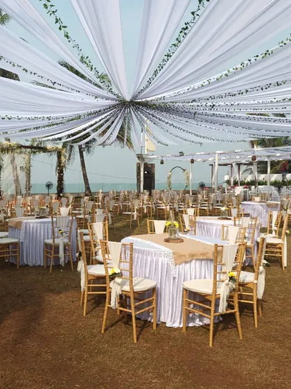 For this rustic beach wedding, we created a beautiful canopy using white drapes and delicate green vines. The guest tables are set with burlap runners, completing the charming and natural aesthetic.