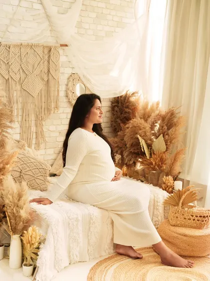 A peaceful portrait of a mother-to-be in a comfortable white dress. She is seated on a bed in a boho-chic room, looking off to the side with a calm and happy expression.