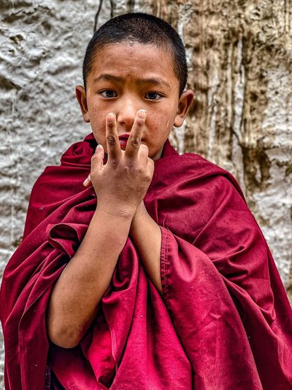 A young monk in Spiti Valley looks at the camera with a curious expression. His bright red robes contrast with the stark, rocky background, making for a compelling portrait.