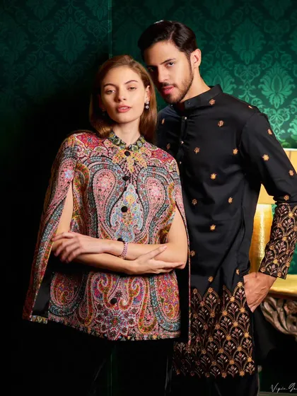 A couple's lookbook shot for an ethnic wear brand. The models are posed against a dark green, textured wall, which makes the intricate embroidery and vibrant colors of their outfits stand out.