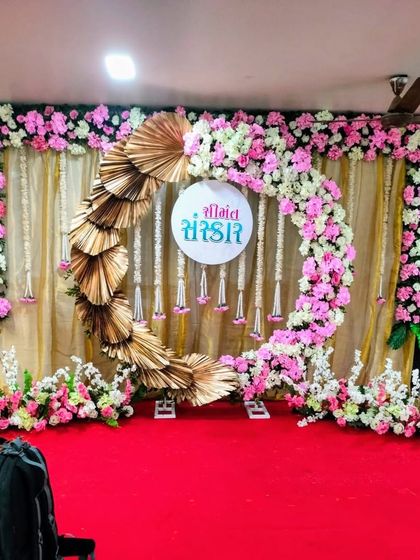 A modern take on a traditional backdrop. This stage for a Sreemantham ceremony uses a circular floral arch of pink and white roses, accented with golden palm leaves against a simple gold curtain, blending contemporary design with classic elements.