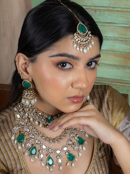 A close-up of the emerald green layered Kundan bridal set, focusing on the maang tikka and jhumka earrings. The combination of uncut stones and pearls creates a look of timeless beauty.
