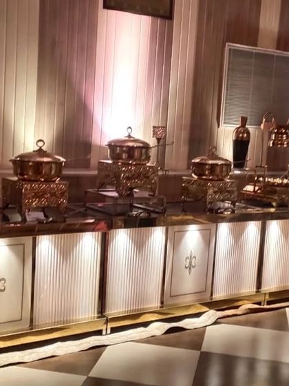 A classic and elegant buffet setup featuring white and gold counters with warm backlighting. The copper serving pots add a traditional touch to the presentation.