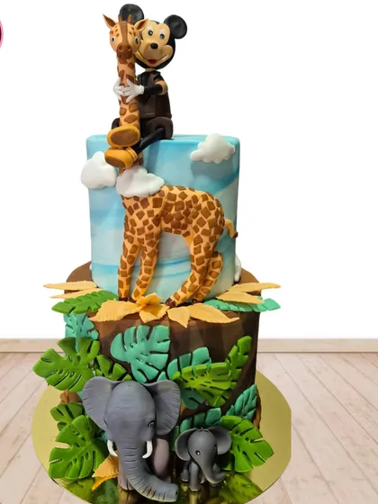 A two-tiered jungle adventure cake featuring Mickey Mouse as a safari explorer. The cake is adorned with fondant elephants, a giraffe, and lush green leaves.