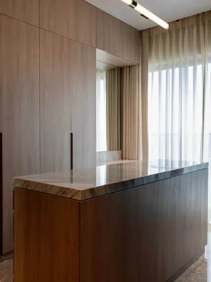 This image captures the serene atmosphere of a custom walk-in closet, featuring a marble-topped island for folding clothes and wood-paneled wardrobes. The large window and sheer curtains fill the space with soft, natural light.