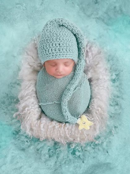Swaddled in teal and wearing a matching pixie hat, this baby sleeps in a fluffy nest against a textured teal background.