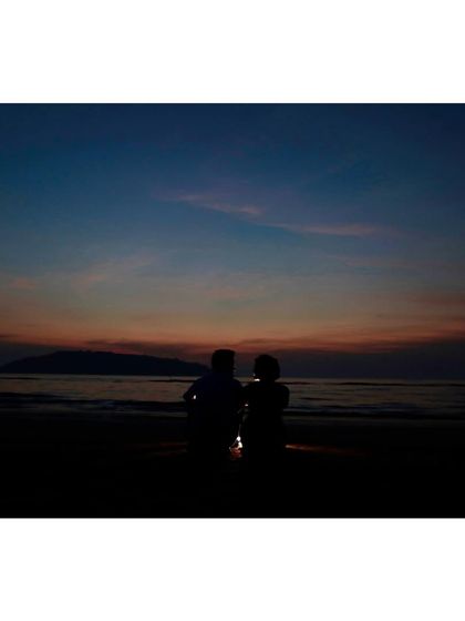A serene silhouette of a couple sitting together on the beach, watching the sunset. This quiet, peaceful moment is perfect for capturing the calm before the wedding festivities.