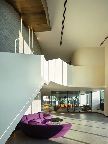 The main lobby is a double-height space designed to feel expansive and welcoming. The bold purple sofa adds a vibrant pop of color against the neutral palette, while the unique angles of the mezzanine create visual interest.