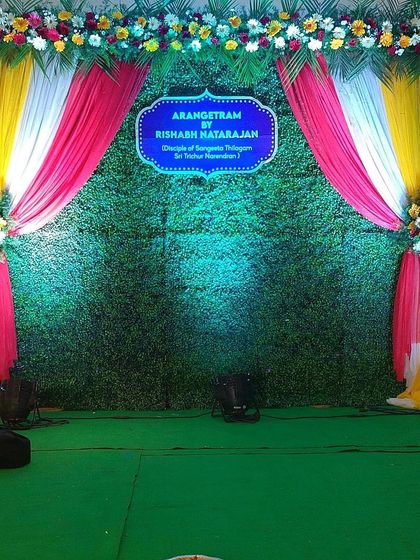This stage backdrop was designed for an Arangetram performance. It features a green turf wall with colorful drapes and floral arrangements, creating a vibrant setting for the dancer.