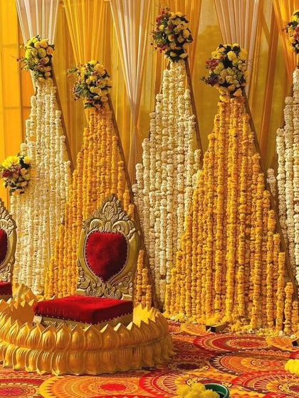 A grand Haldi stage with a backdrop of marigold and tuberose strings arranged in triangular patterns. The two ornate golden chairs with red cushions add a royal touch to the ceremony.