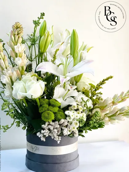 An elegant and serene floral arrangement in a grey velvet hatbox. This stunning bouquet features white lilies, tuberoses, and roses, creating a classic and timeless look of pure sophistication.