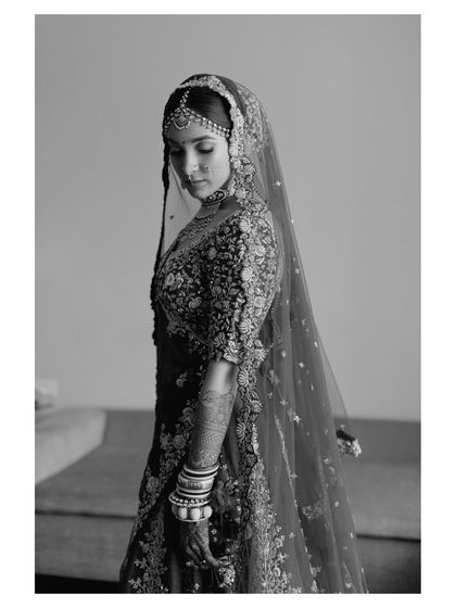 A black and white version of a classic bridal portrait. The lack of color emphasizes the intricate embroidery of her lehenga and the timeless beauty of her pose.