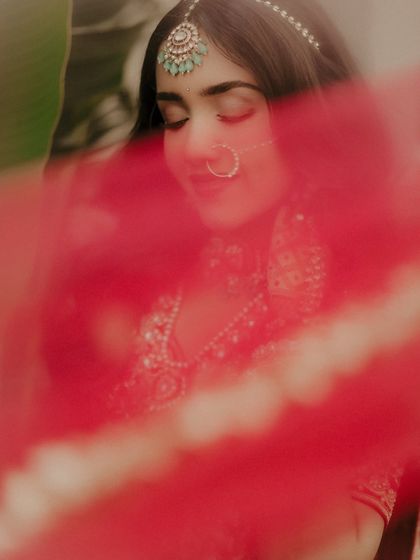 An artistic shot using the bride's dupatta. The sheer red fabric creates a beautiful, soft veil over the frame, adding a layer of mystery and romance to this close-up portrait.