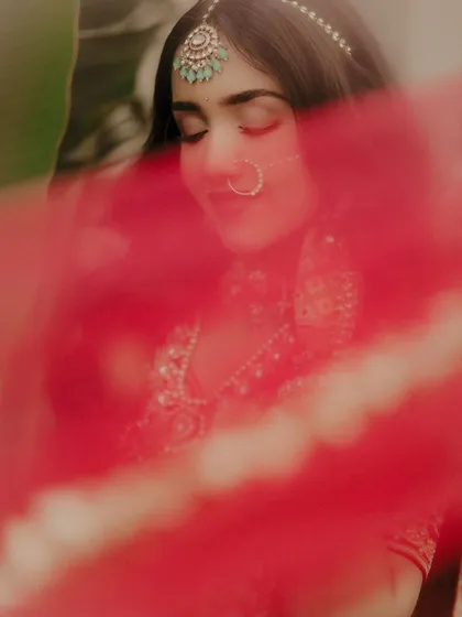 An artistic shot using the bride's dupatta. The sheer red fabric creates a beautiful, soft veil over the frame, adding a layer of mystery and romance to this close-up portrait.