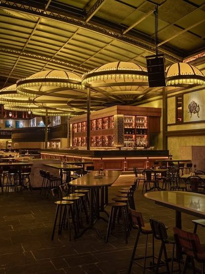 A wide shot of the main floor, showing the relationship between the bar, high-top tables, and the surrounding architecture. The open layout is designed to feel energetic and communal.